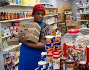 A woman of colour stands inside of a grocery store. She is wearing a blue apron, signifying she is an employee there. She is holding four boxes of fruit in her arms.