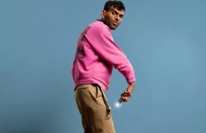Comedian Aaqib Merchant poses in front of a blue background. He is wearing a pink sweater.