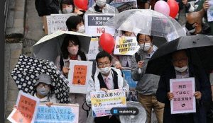 A crowd of Japanese citizens gather together in the rain to protest. On is holding a sign that reads "Long Detention is torture!"